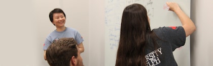 Student writes on white board while professor watches and smiles
