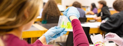 Student wearing blue latex gloves holds a small plastic package of green liquid and takes samle