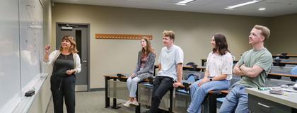 4 Students sitting on desk in a classroom watching professor as she writes on the whiteboard.