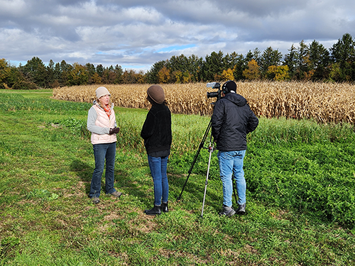 Dr. Laure Van Eerd standing in a field while being video recorded by two people