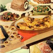 Cookbook cover with roast chickens, veggies, tarts, and dessert roll on a table.