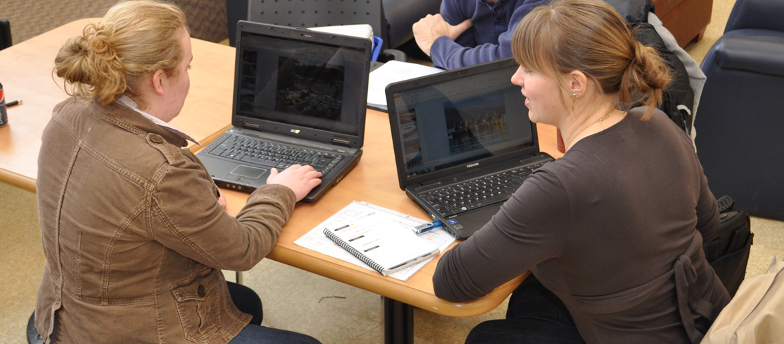 Two female students sit with laptops, backs to the camera