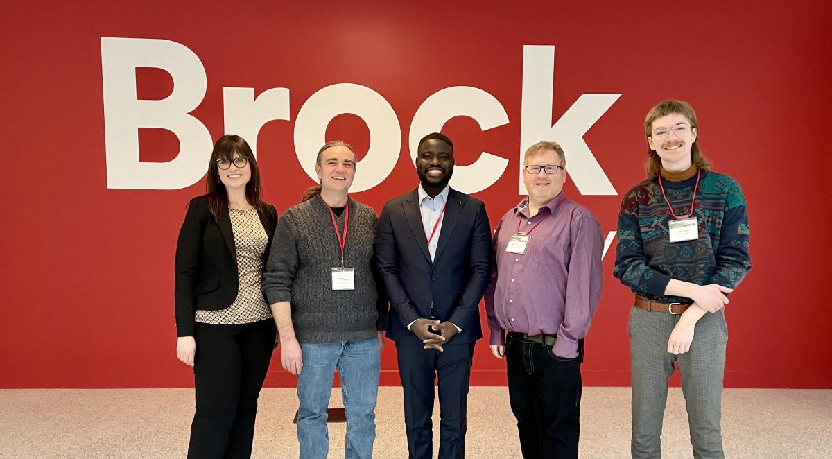Five people standing in front of University of Brock red and white wall