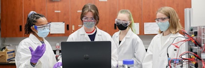 Four co-op students wear lab coats, medical masks, and medical gloves, gathered around a computer discussing.