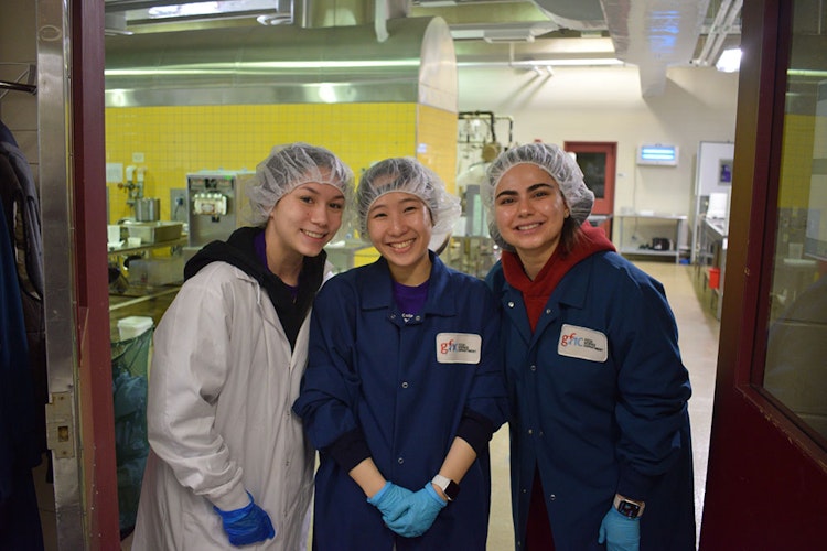 Three students in lab coats and hair nets smile in a food science lab.