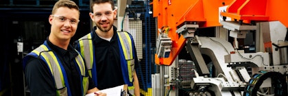 Two people wearing high visability vests smile at the camera, standing beside engineering automotive equipment.