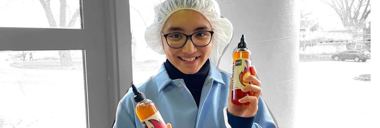 A student wearing glasses, a lab coat and hair net holds up two bottles of hot honey.