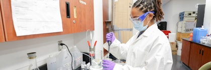 Person wearing safety goggles, medical mask, lab coat, and medical gloves uses a pipette in a lab setting.