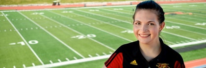A person smiles at the camera wearing a red and black shirt, in front of a sports field.