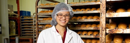 Person wearing a lab coat and a hair net smiles, standing beside bakers' racks of bread.