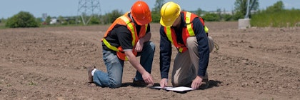 Two people wearing construction vests and hard hats kneel on dirt ground outside, looking at a paper.