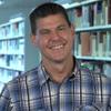 Trevor smiles in blue plaid shirt in front of bookcases