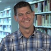 Trevor smiles in blue plaid shirt in front of bookcases