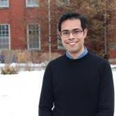 Nuno stands outside with a snowy building behind him, smiling in a collared shirt, sweater and glasses
