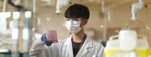 Student in a lab coat and mask standing at a lab desk holding a stack of petri dishes and lab equipment around him.