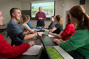 Students at table with laptops look to prof speaking at front of room