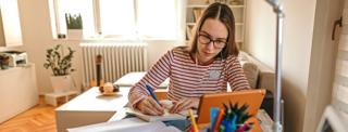 Female sitting at a desk, writing in notebook while looking at the tablet screen in front of her
