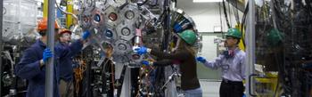 four people wearing hard hats working on a large piece of equipment in a big lab
