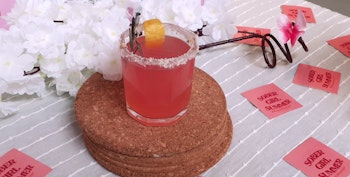 A pink drink with sugar rim and lemon zest on a cork coaster, surrounded by white flowers and 'Sober Girl Summer' cards.
