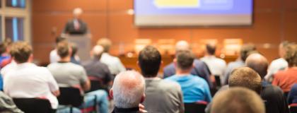 A presenter stands at the podium in front of a crowded lecture hall. Presentation slides are projected on a screen behind them.