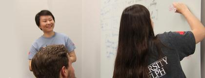 Student writes on white board while professor watches and smiles
