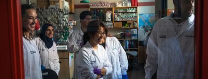 View through a red door, group of students in lab coats smile toward professor in food lab