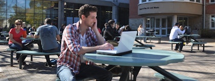Picture of student sitting at a picnic table outside while working on a laptop. 