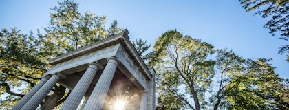 Photograph of the Portico with sun rising, peeking through the trees behind it. 