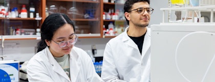 Two students working in lab, one male/one female wearing lab coats and glasses