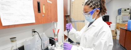Person wearing safety goggles, medical mask, lab coat, and medical gloves uses a pipette in a lab setting.