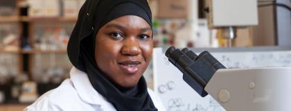 Person smiles at the camera, in front of a microscope in a lab setting.