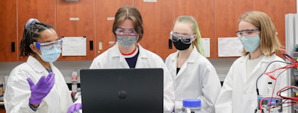 Four co-op students wear lab coats, medical masks, and medical gloves, gathered around a computer discussing.