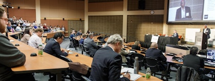The 2019 Gryphon's LAAIR event hosted in a large lecture hall with tiered seating. Attendees, some taking notes, are focused on a presenter speaking at the podium. The speaker is also displayed on a large projection screen behind him.