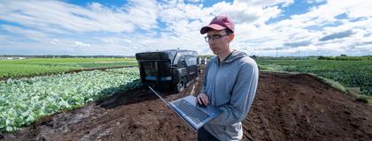 Person holding laptop while standing in a crop field with a large robotic weeder in the background.