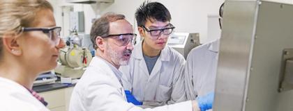 A group of individuals in lab coats examining an object with focused expressions in a lab.