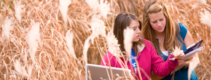 two female students in a wheat field, holding laptops, inspecting the crops
