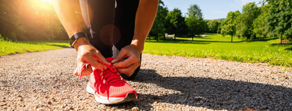 A person tying the laces of a bright pink running shoe on a gravel path in a park surrounded by green trees and grass under a sunny sky.