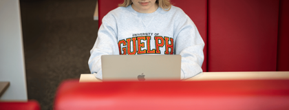 Student wearing a Guelph sweater sits in a red booth typing on their laptop.