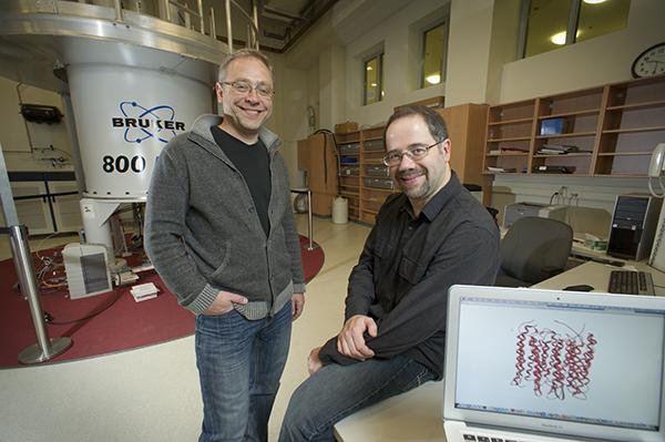 two men, one seated on standing in a lab with large equipment in the background and a computer screen in the foreground