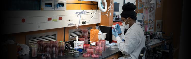 Student in a lab coat and mask sitting at a lab desk working on piping.