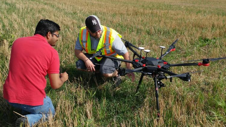 Two people examining a drone in a field. One is wearing a red shirt, and the other is in a reflective vest. The drone has multiple rotors and is positioned on the ground with tall grass and crops around.