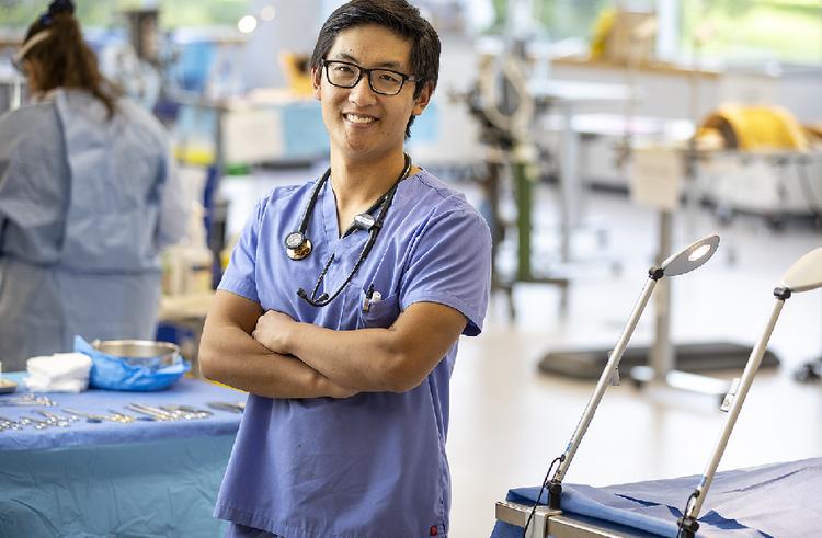Student in medical scrubs within a laboratory environment. Arms crossed, smiling at the camera.