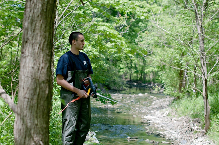 person in the forest holding one end of a measuring tape across a stream