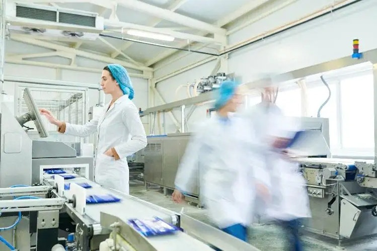 People in lab coats and hair nets walk by clean assembly line machinery