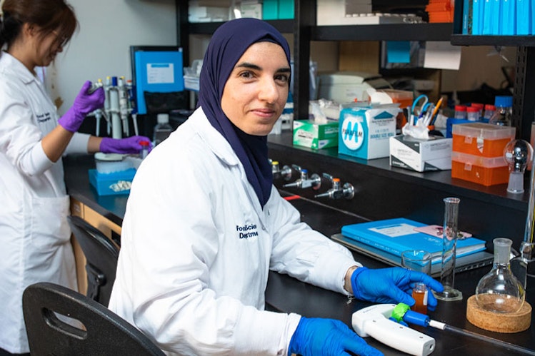 Female student in lab coat and hijab smiles at lab work bench
