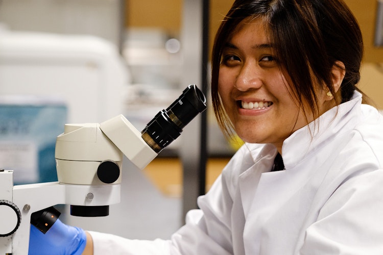 Person with side bangs smiling in front of microscope. In foreground and background are blurry lab materials.