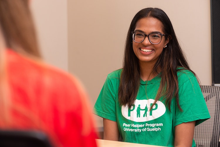 Person with long dark hair and glasses, wearing a green Peer Helper Program tshirt, smiles at someone in front of them.