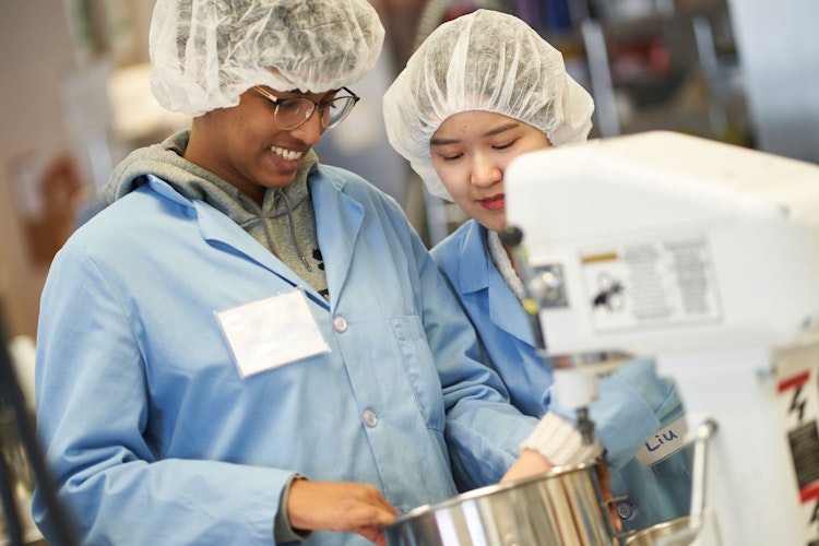 Two students in hair nets and blue lab jackets look down into stand mixer bowl