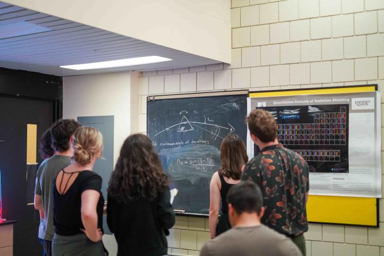 Students standing at a chalkboard looking at a math problem.