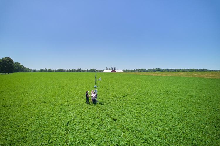 Aerial view of two people standing in a vast green field with weather monitoring equipment, with a distant farmhouse and blue sky overhead.